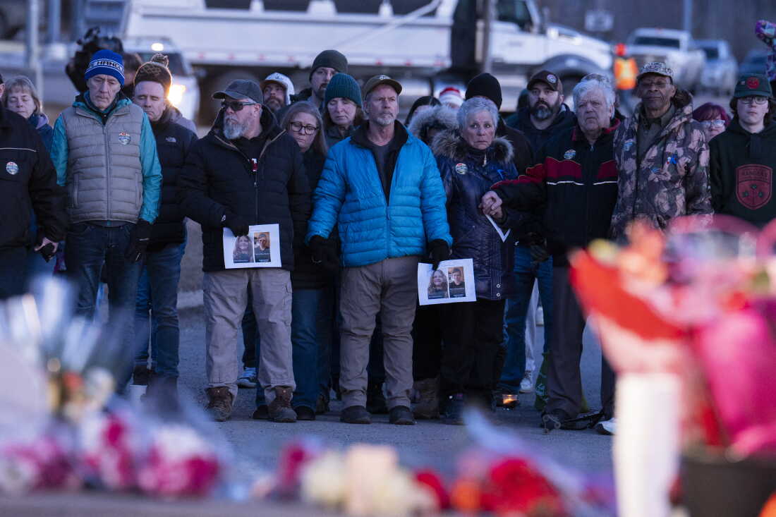 Community members stand shoulder to shoulder, some holding hands, at a vigil. There are bouquets of flowers on the ground.