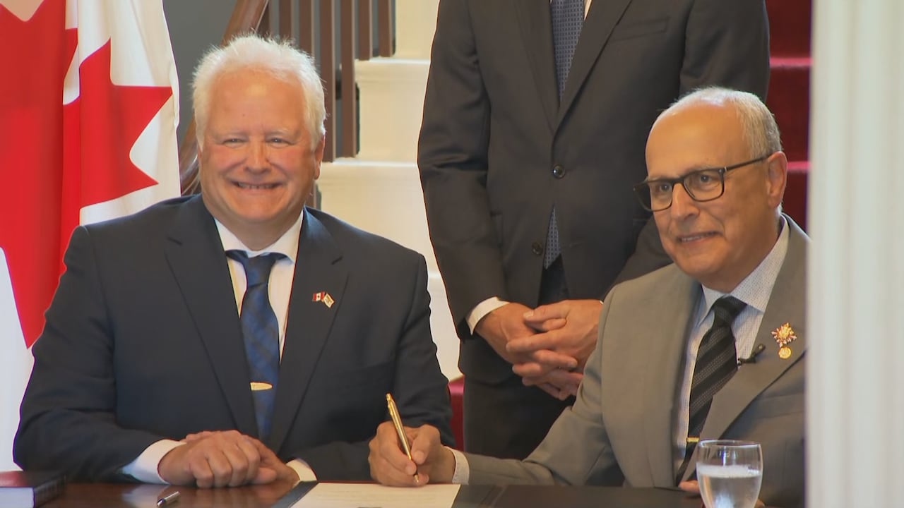 Two men sit at a formal table after signing a document. They are smiling at a camera.