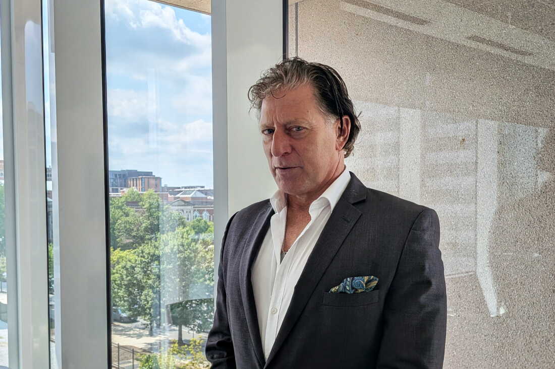 Richardson is wearing a dark suit and a partially unbuttoned white shirt.