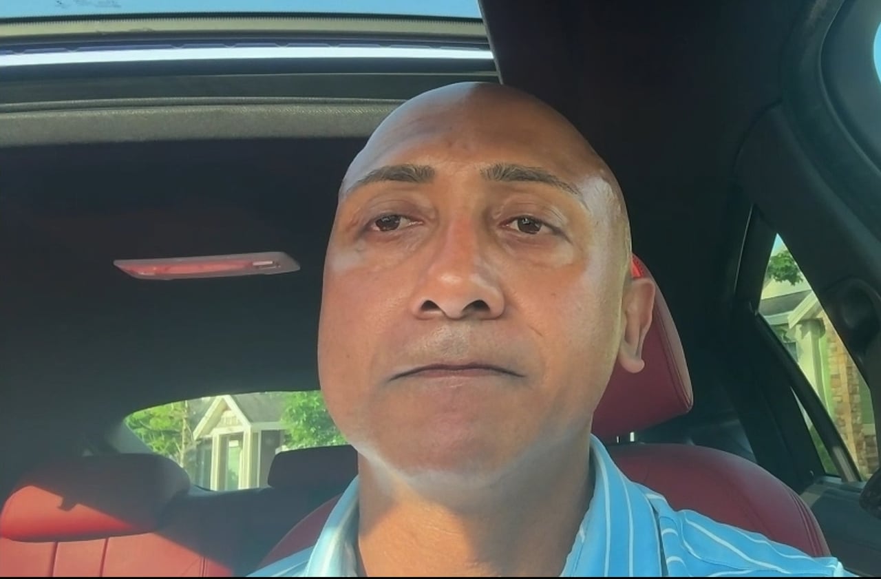 A bald South Asian man speaks in a car.