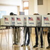 Voters in Detroit stand in voting booths that say "Vote" with an American flag on the side. We can see their legs and feet but not their faces, which are obscured by the voting booth.