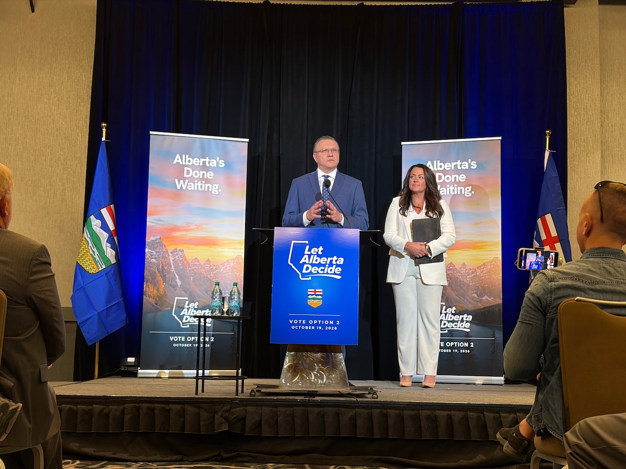 A man and woman stand on a small stage at a podium. Behind them are banners that say 'Alberta's done waiting.'