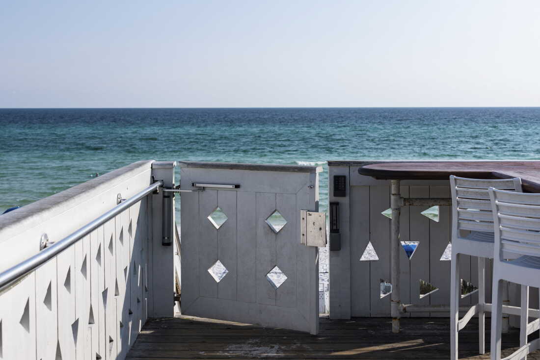 A gray wooden gate on the edge of a wooden beachside deck is equipped with a keypad to grant access to the beach behind it, near the Coleman Pavilion obelisk in Seaside, Florida. Blue waters are in the background.