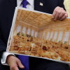 President Trump holds papers that show a rendering of his proposed White House ballroom. He is wearing a suit with a blue tie and is sitting in an armchair.