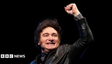 Argentine President Javier Milei, wearing a leather jacket, stands smiling with a closed fist raised against a black backdrop.