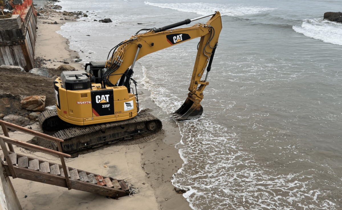 A photo of an excavator on a narrow strip of beach.