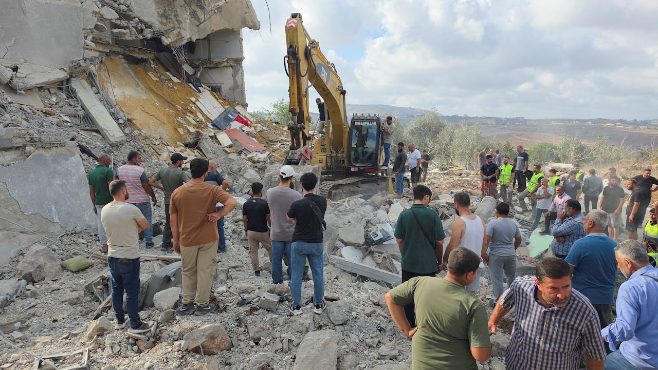 People are seen standing next to an excavator by a destroyed building.