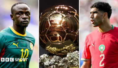 A triptych showing Sadio Mane, the Ballon d'Or trophy and Achraf Hakimi. Mane wears a green Senegal kit with a yellow number 10 on the chest while Hakimi wears a red Morocco shirt with a gold number two on the chest. The golden Ballon d'Or trophy which sits between them is a football divided into hexagonal patches sitting atop a glittering chunk of mineral ore