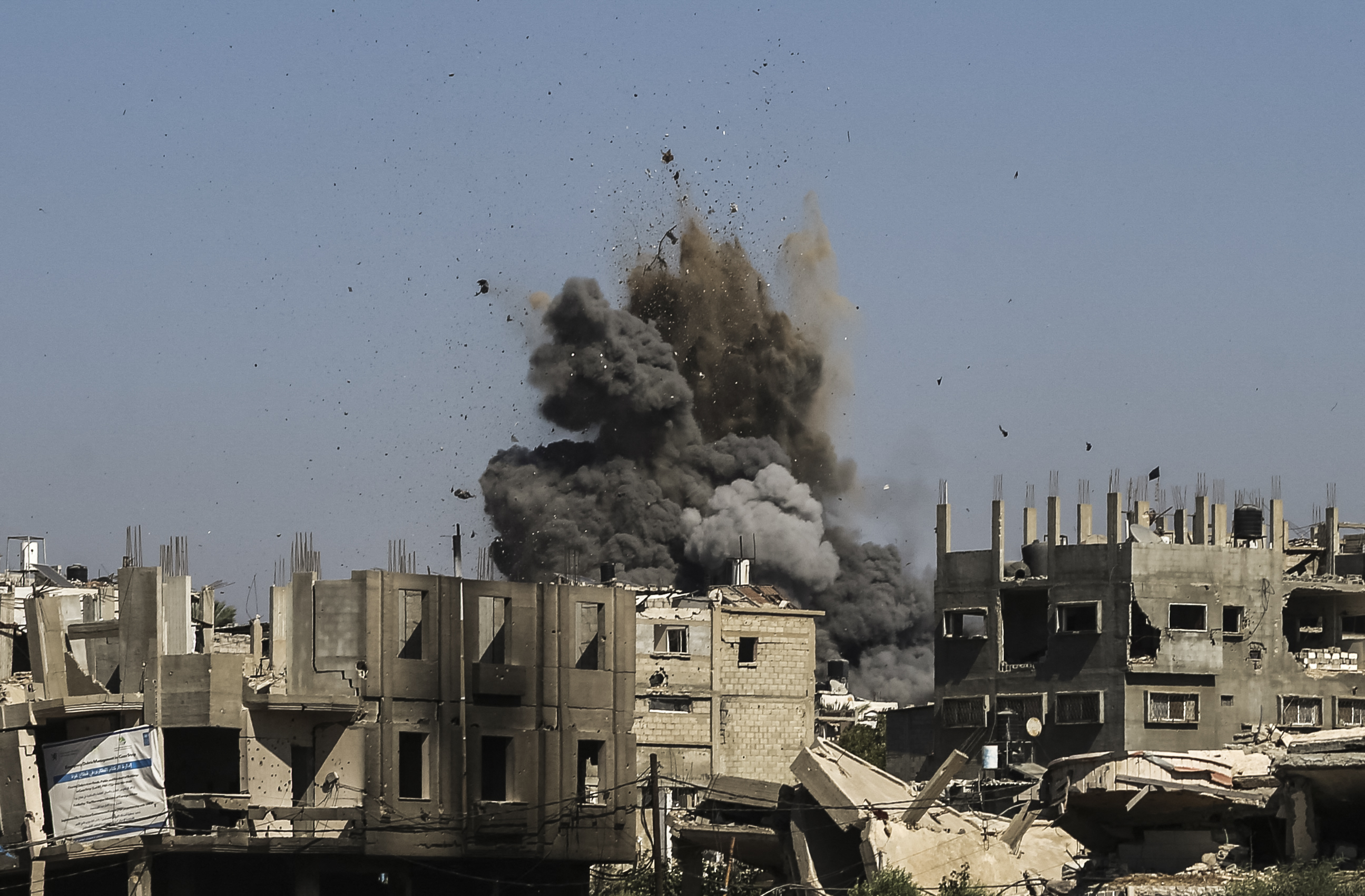 A large explosion erupts in a war-torn area, sending a cloud of smoke and debris into the air as buildings lie in ruins from ongoing conflict.