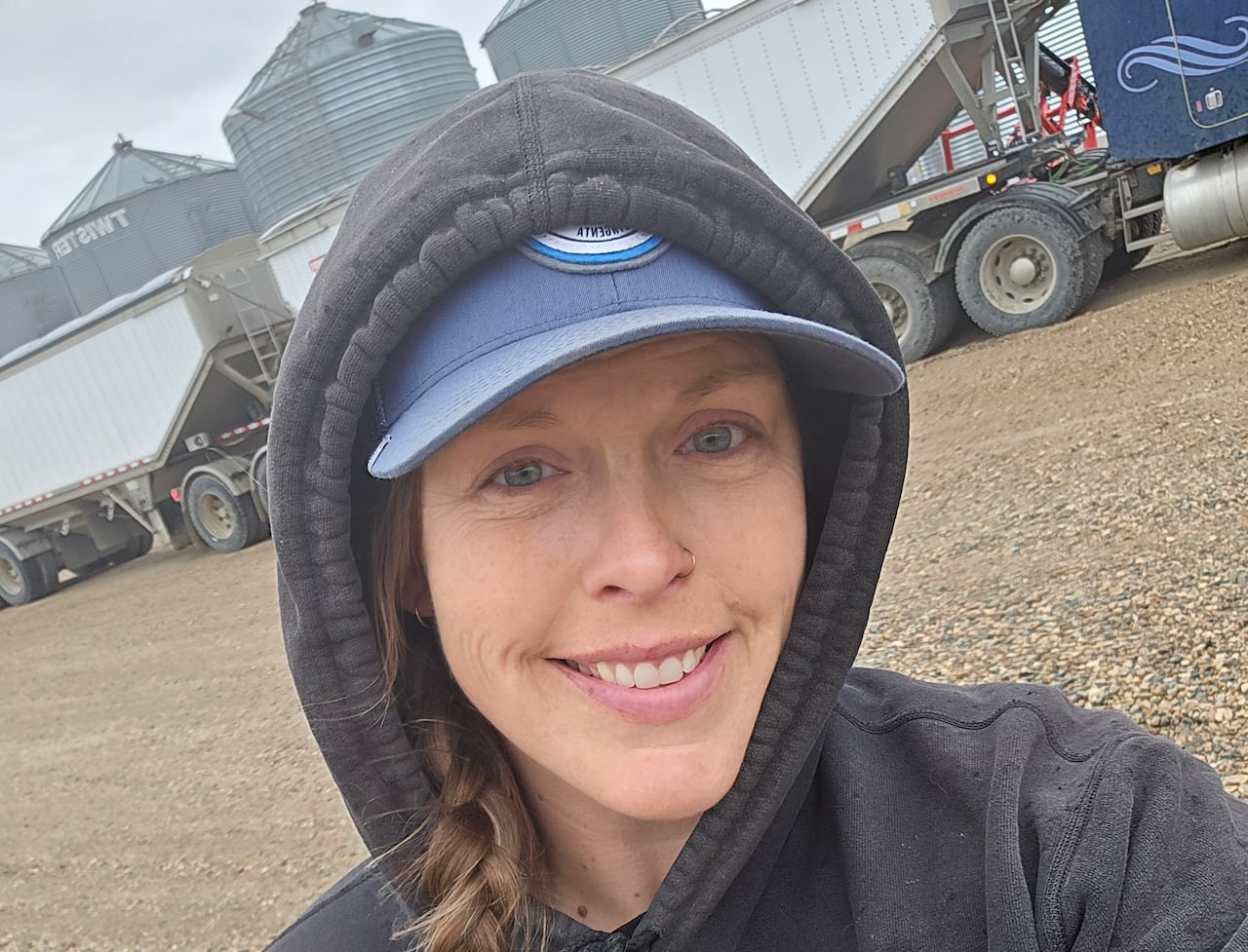 A woman with a braid and in a dark grey hoodie smiles at the camera. A large truck and grain bins are in the background.