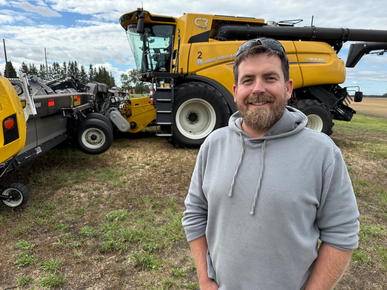 A man in a grey sweater smiles at the camera and stands in front of farm equipment.