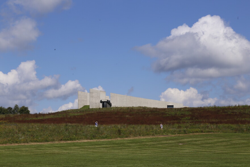 The concrete visitor center at Flight 93 National Memorial sits atop a grassy hillside as two visitors walk below under a blue sky filled with large white clouds.