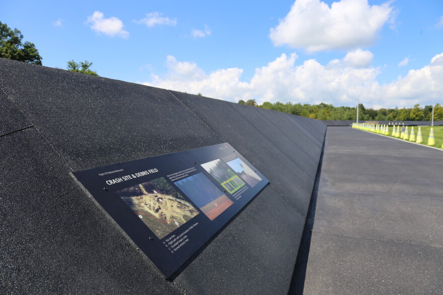 An interpretive panel labeled “Crash Site & Debris Field” is mounted along a dark memorial wall beside a paved walkway under a bright blue, partly cloudy sky.