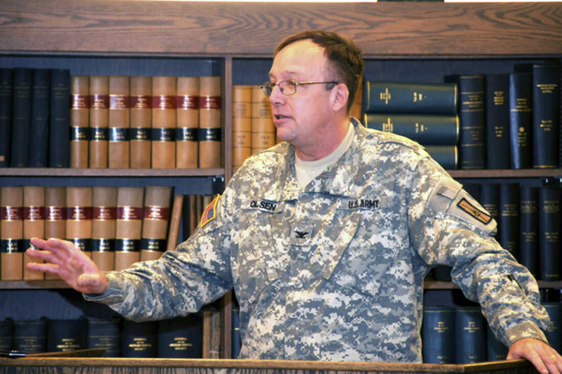 In this photo, the Rev. Eric Olsen is wearing a camouflage Army uniform with a pixelated pattern. Behind him are bookcases with rows of books.