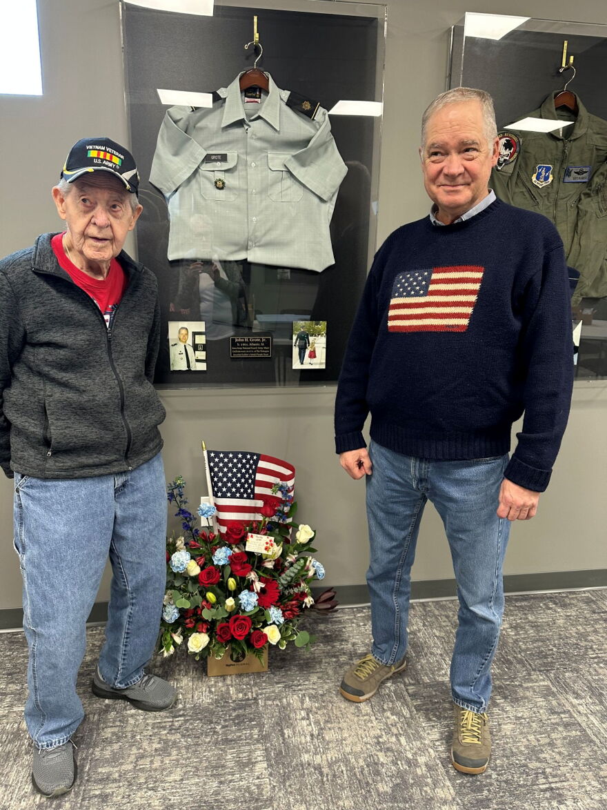 Two men stand in front of a display featuring a military uniform. There is a flower arrangement between them on the ground.