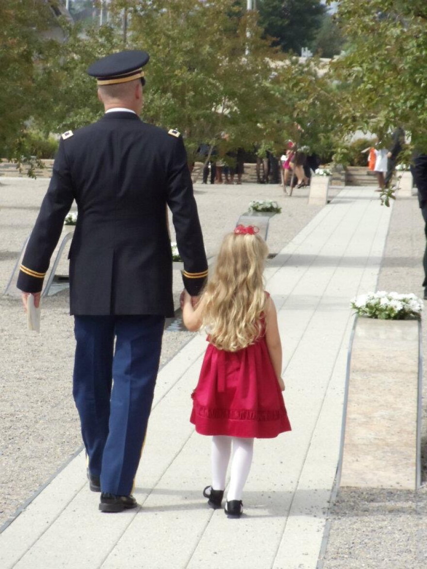A soldier walks hand-in-hand with a young girl with long blonde hair and a red dress.