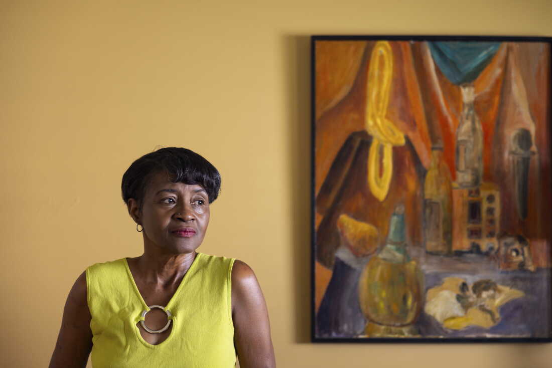 Allie Zemtseff stands in front of a yellow wall with a painting hanging on it. She's wearing a yellow sleeveless top.