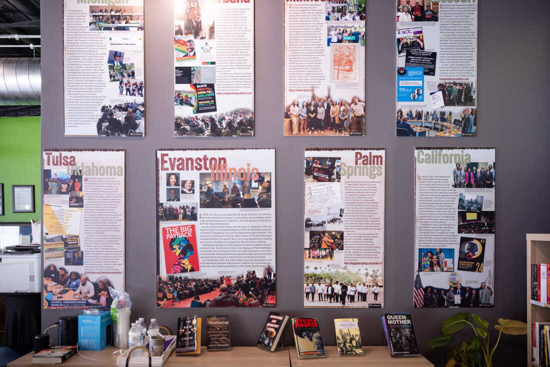 Explainer posters hang on an office wall at the nonprofit FirstRepair in Evanston, Illinois. Each poster highlights local reparations efforts in a specific U.S. location, such as Tulsa, Oklahoma, and Palm Springs, California.