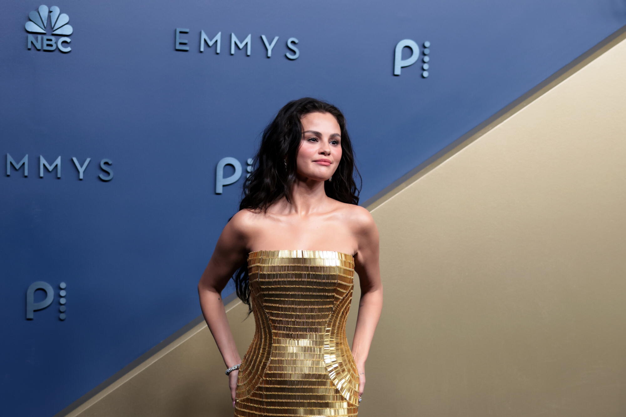 Selena Gomez, wearing a gold gown, poses on the Emmys blue carpet. 