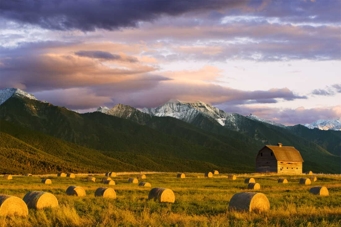A field of hay bales frame an old western wooden barn with the dramatic Mission Mountain Range in the background near Pablo, Montana.