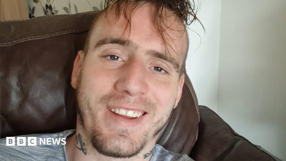 William Watson looking straight at the camera. He is sitting in an armchair. He is smiling with his teeth on show. He has long dark hair over his forehead and a short, dark beard. He has tatoos on either side of his neck and appears to be wearing a grey t-shirt.