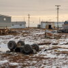 A shed-like building and an all-terrain vehicle lie overturned upside down in Kwigillingok, Alaska, in late October. The ground is partially covered by snow. In the background stand a few small, one-story buildings. Power lines supported by poles crisscross the area.