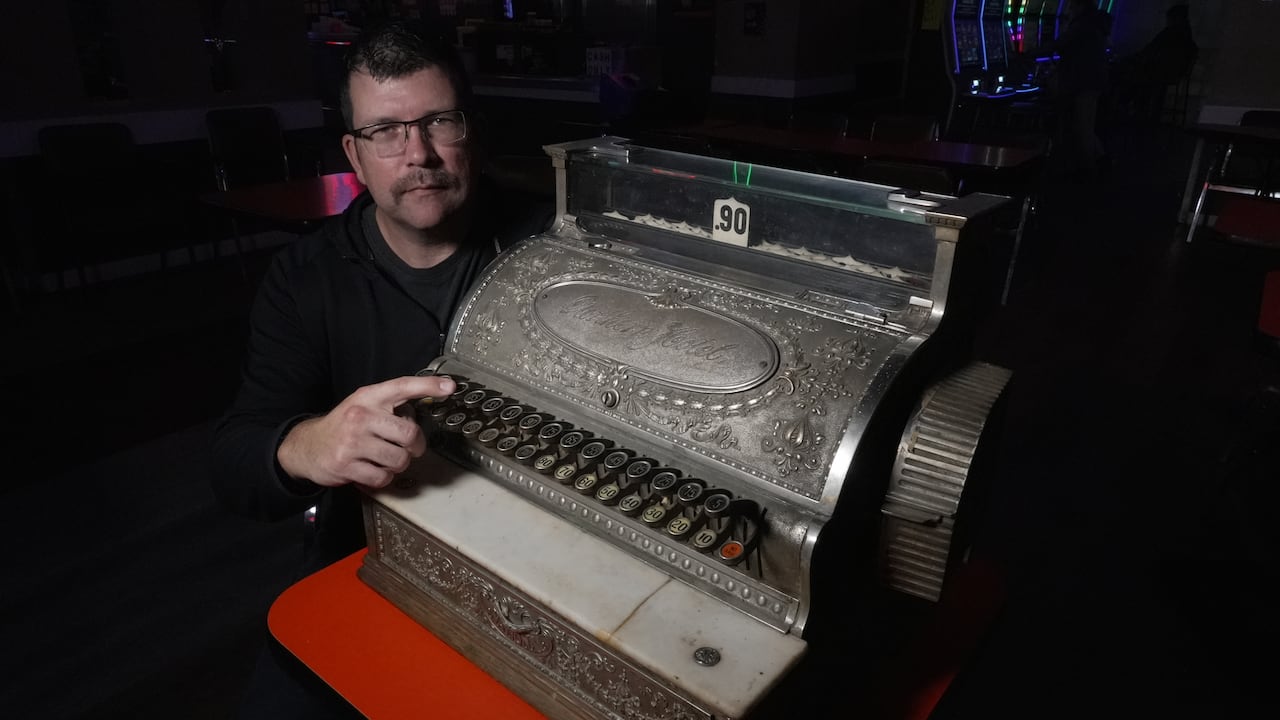 A man sits behind a large mechanical cash register with his finger on one of its buttons.