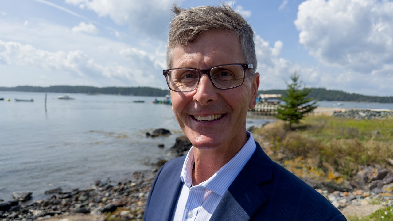 A smiling man with glasses in a blue blazer and button down shirt stands in front of a harbour with with wharf, water and trees in the background.