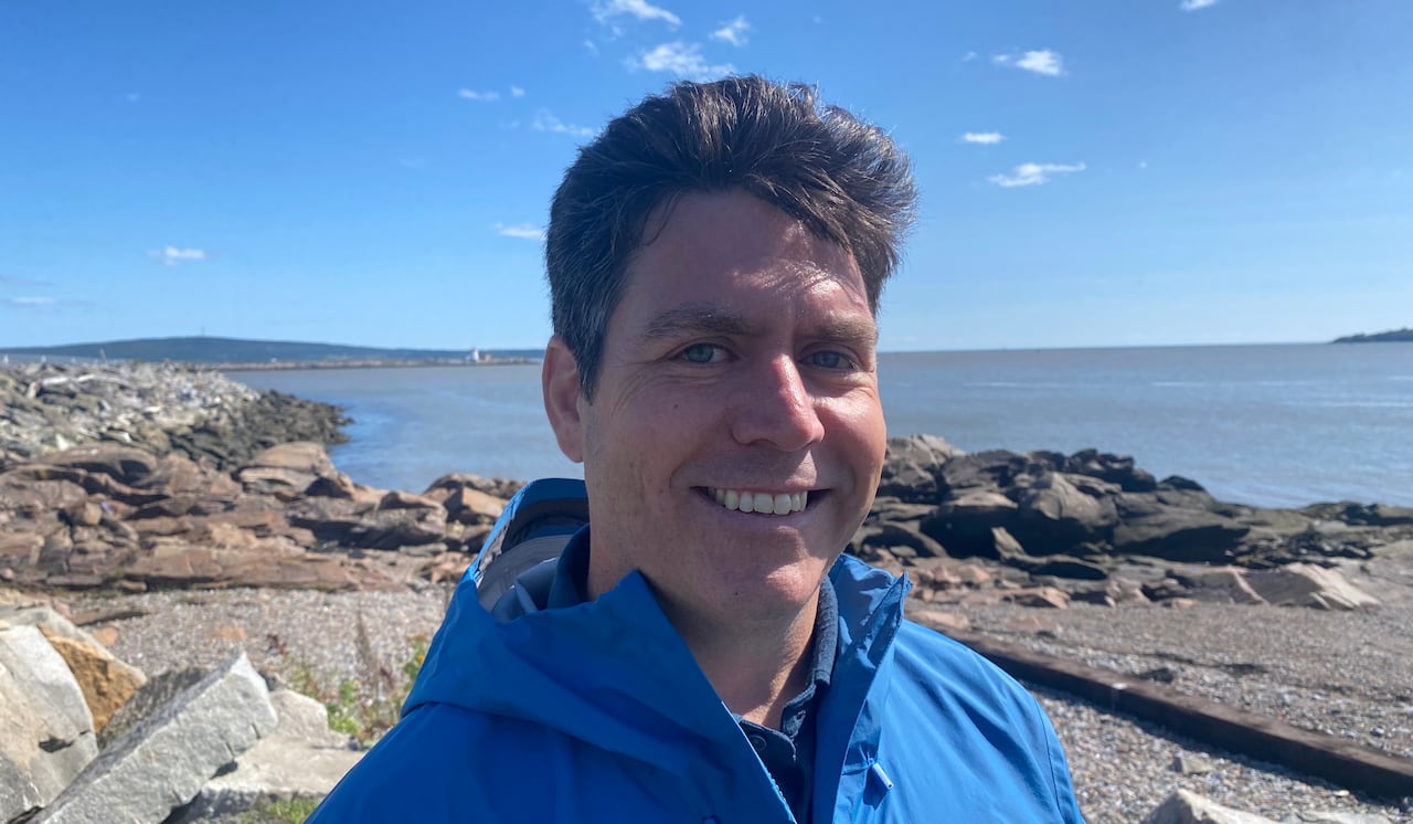 A smiling man in a blue coat stands in the shore of a harbour with the water and ski in the background.
