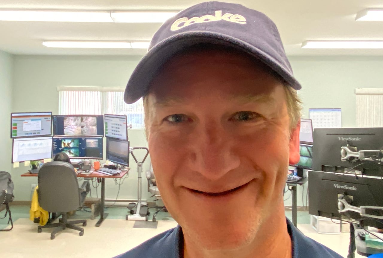 A smiling man in a blue ball cap stands in front of a woman working at a computer in a room.