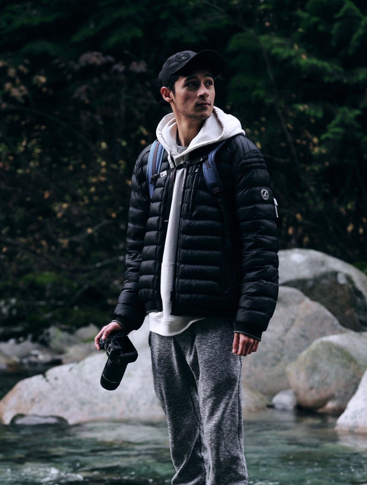 A man stands in front of a stream holding a camera