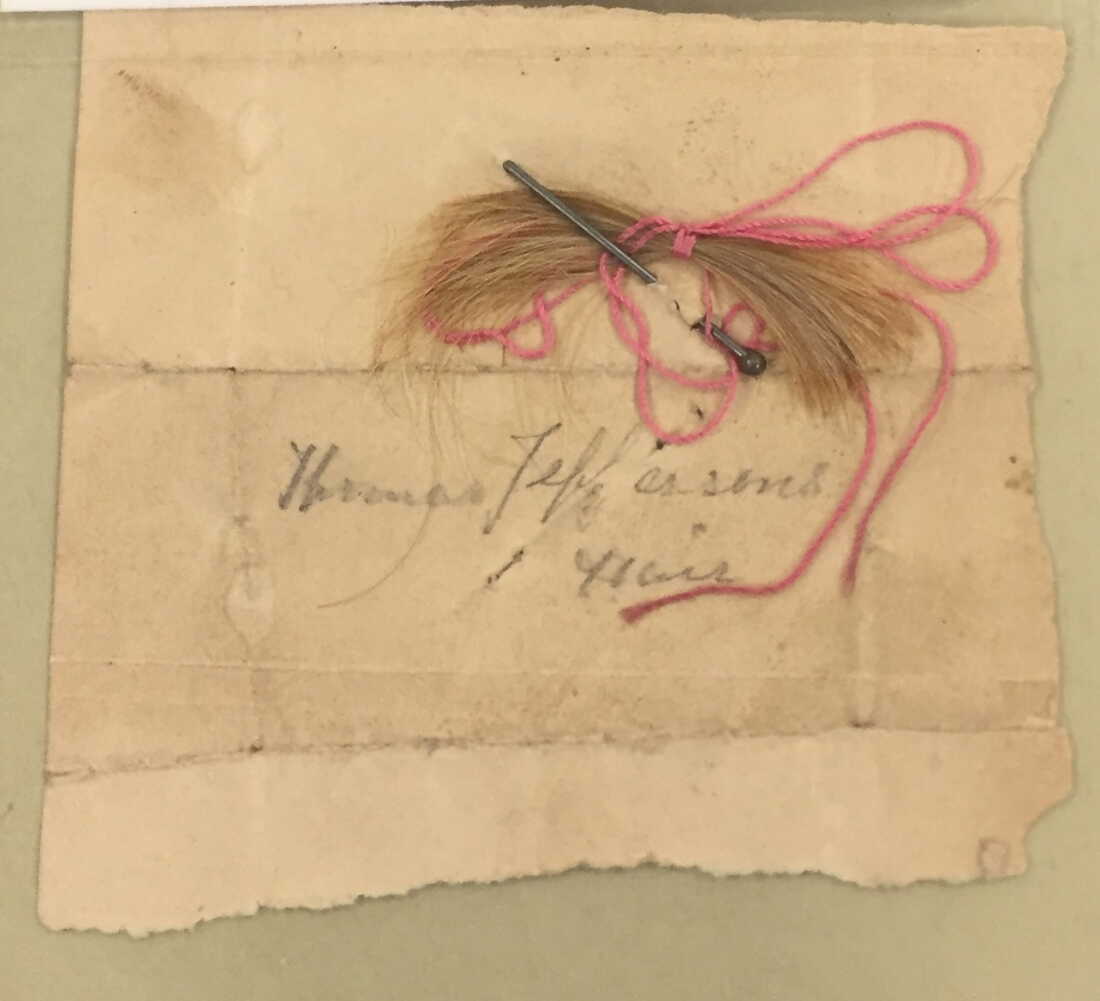 A clipping of hair labeled as Thomas Jefferson's is tied together in the middle with a pink string. The hair is colored blond to light brown. The clipping is lying on an old, yellowed piece of paper on which is written in old-style cursive: "Thomas Jeffersons Hair."