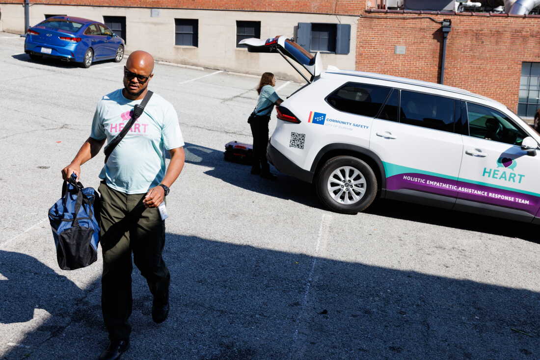 Rob Crisp walks in a parking lot as he responds to a call for a wellness check at the lot. He's carrying a duffel bag. Behind him is an SUV with its rear door raised up; the side of the vehicle has the words "HEART," "Community Safety" and "City of Durham." A woman is reaching into the vehicle's trunk.
