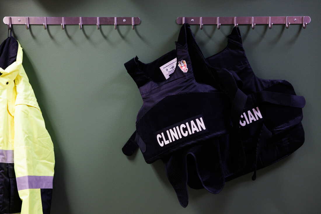 Two black bulletproof vests that say "CLINICIAN" in white capital letters hang on hooks on the wall of the HEART program's office.