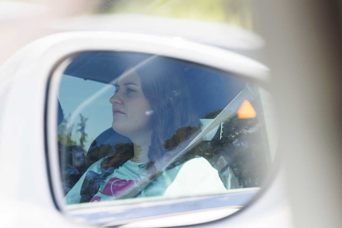 This photo is of the reflection of Allie Craft in the side-view mirror of a motor vehicle. She appears from roughly the shoulders up, and a seat belt spans diagonally across her torso.