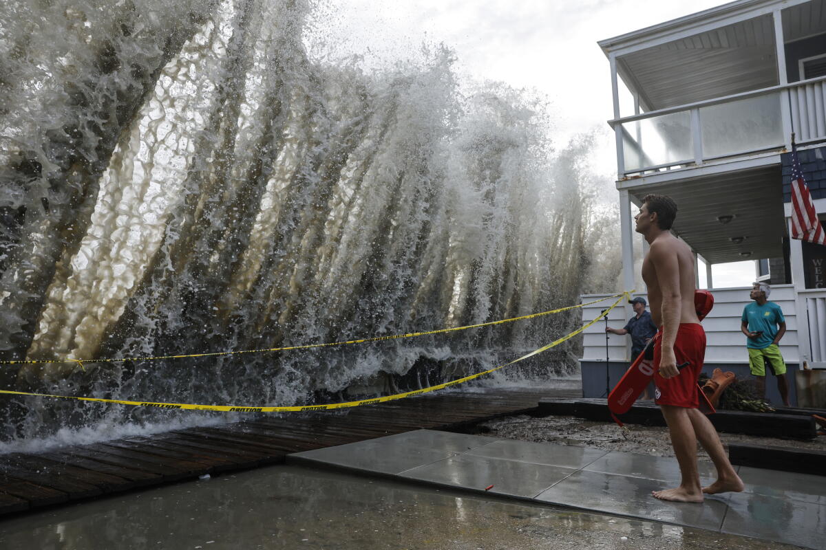 A lifeguard keeps watch along a closed boardwalk as a wave crashes over a seawall.