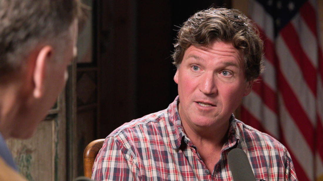 Tucker Carlson is seen from the shoulders up in a plaid collared shirt. He sits in front of a mic.