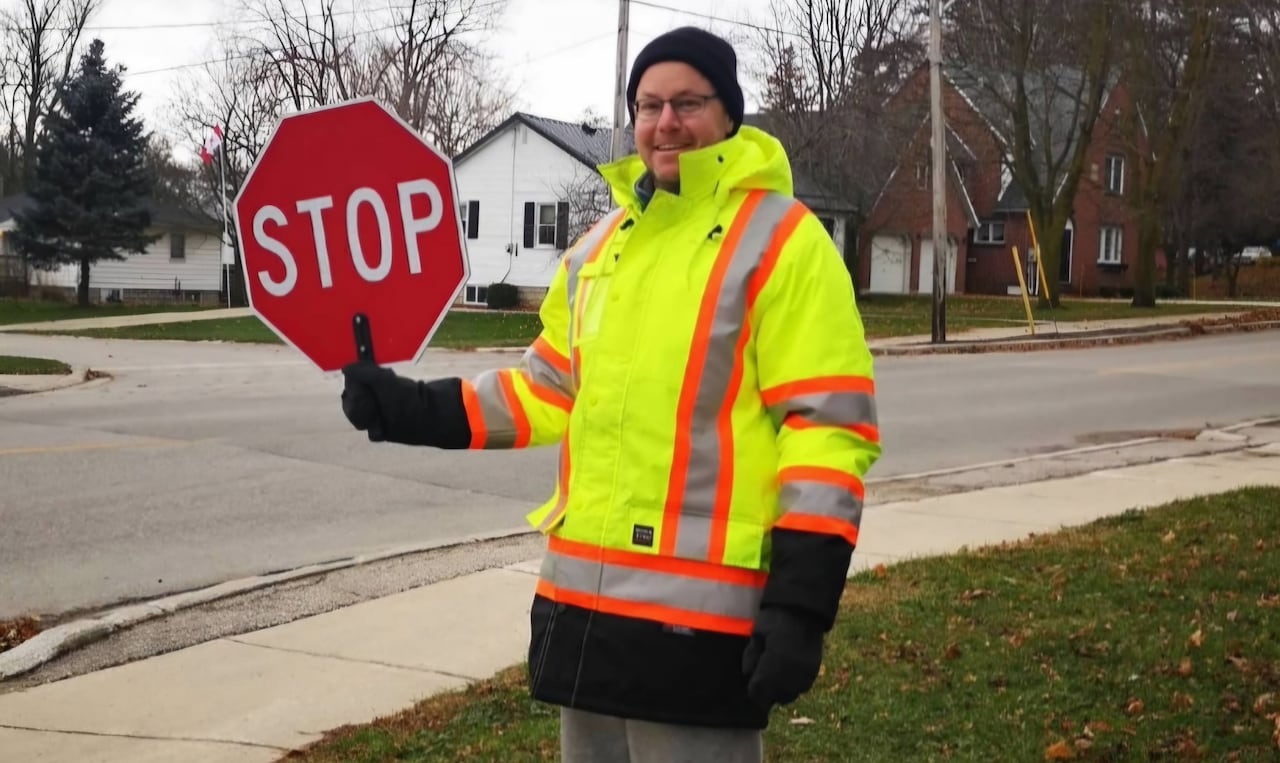 A man in a bright vest holding a stop sign at an intersection, and smiling.