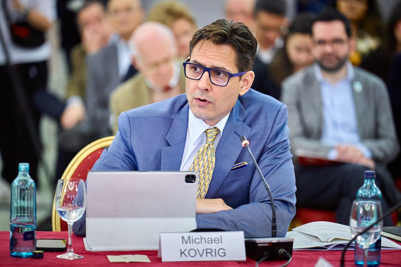 A man in a blue suit and matching glasses sits at a table in front of a tablet, a microphone and a nameplate that reads "Michael Kovrig."