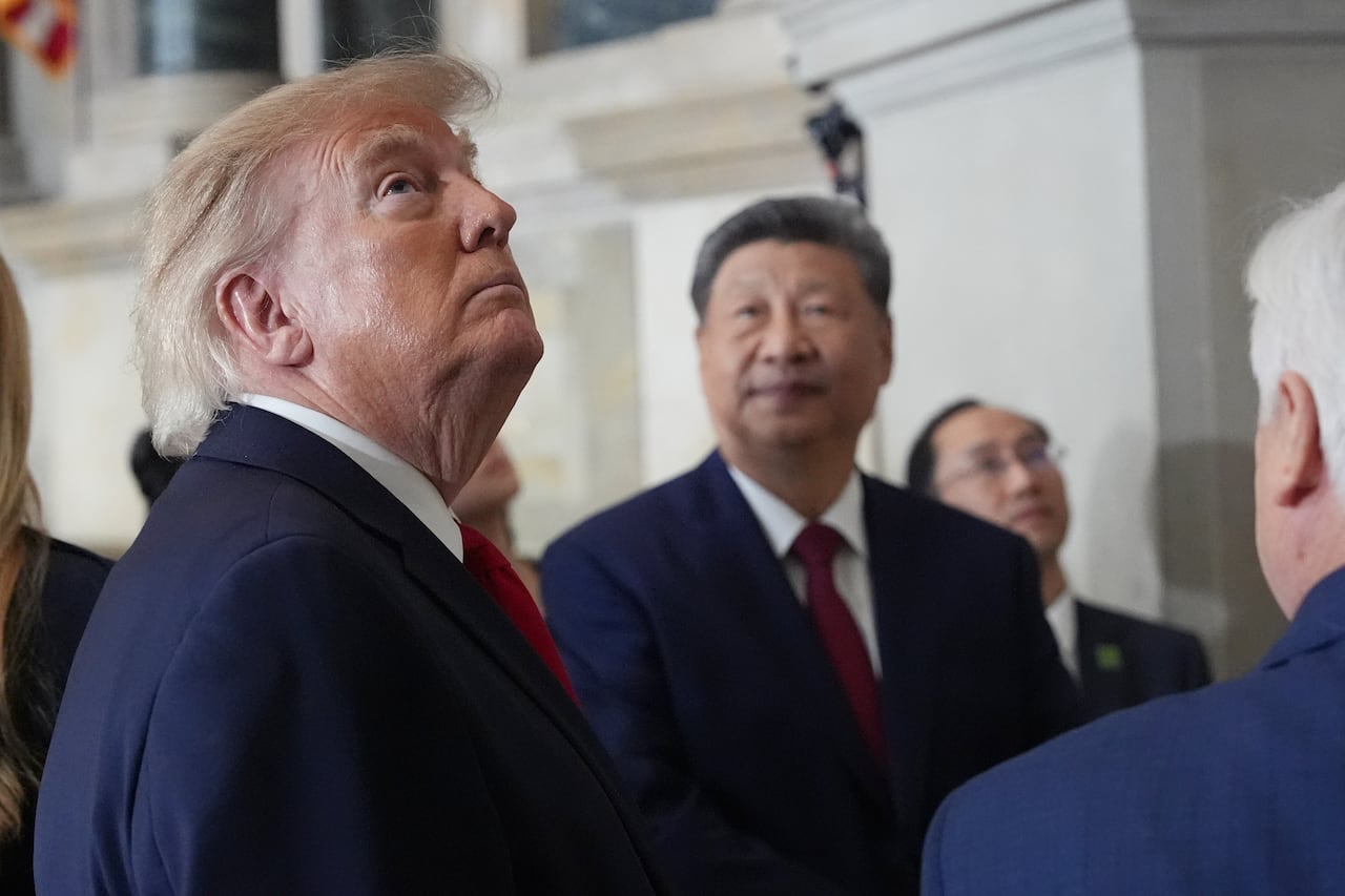 Donald Trump looks upwards while Xi Jinping watches him. Both men are wearing matching dark blue suits and red ties.