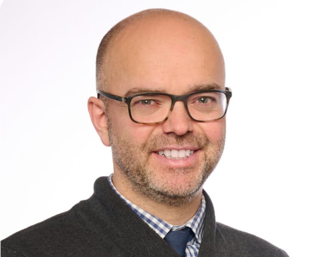 Headshot of a smiling man with glasses, short facial hair, and a dark sweater over a collared shirt against a white background