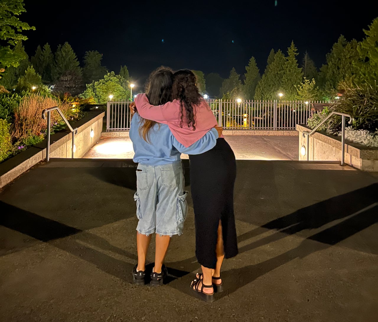 Two women put their arms around each other and look out at a garden lit at nighttime.