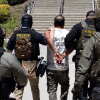 Four Immigration and Customs Enforcement agents walk a detainee out of the Ventura County Government Center in Ventura, California, on July 27. The individuals are photographed with their backs to the camera, and the detainee is handcuffed behind his back. The agents are wearing bulletproof vests that say "POLICE ICE" in yellow capital letters.