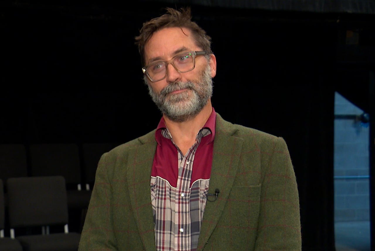 A man in a button up shirt and green blazer looks off camera.