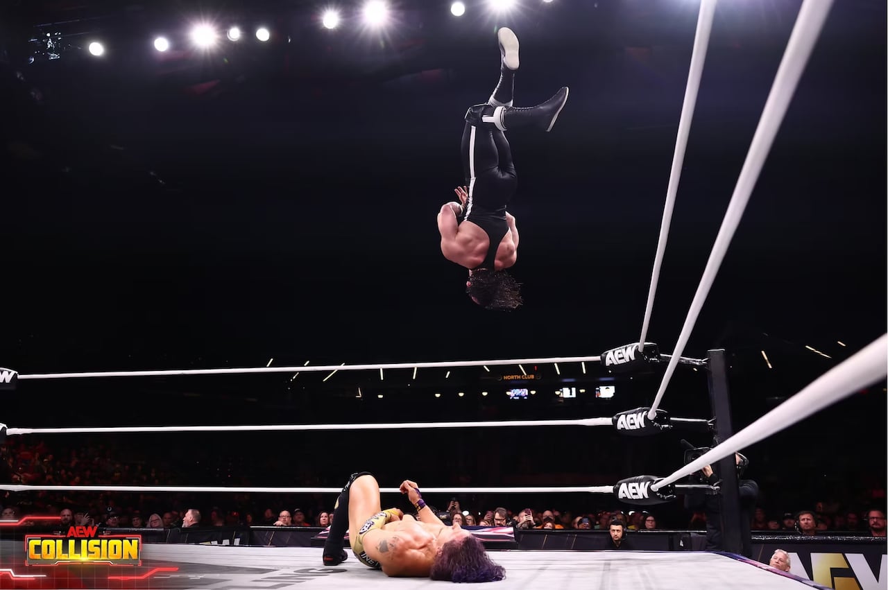 A man in a black wrestling single is in the air above another wrestler prone on the ring mat.