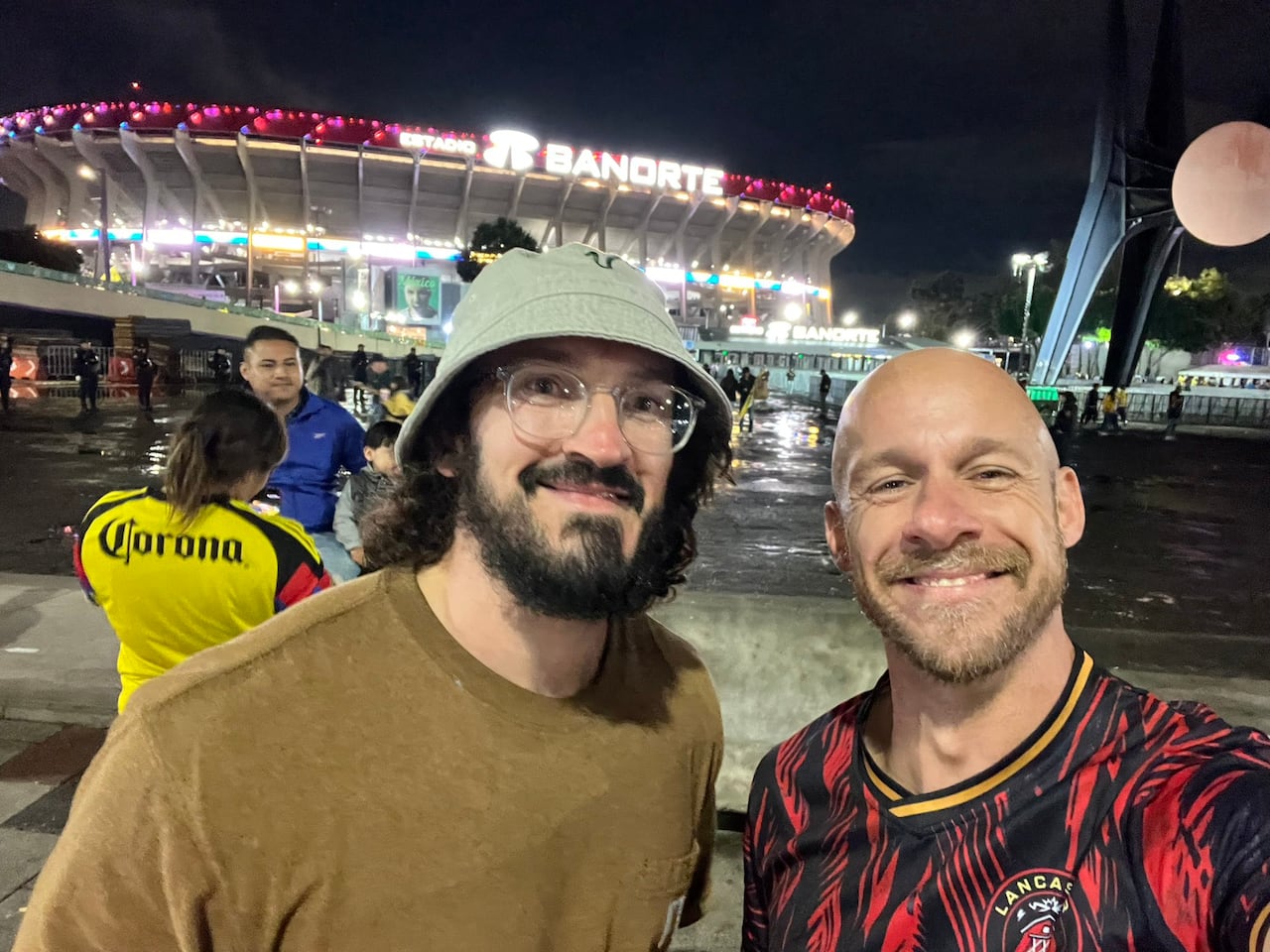 A photo of an adult man with brown hair and bear wearing a bucket hat smiling next to a bald man wearing a black and red football shirt.
