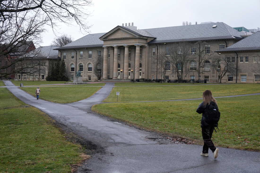 Two women wearing backpacks walk on a paved pathway on the campus of Cornell University in Ithaca, N.Y., in 2024. The pathway cuts through an open area with green grass. In the background is an old-style, three-story building with four pillars.
