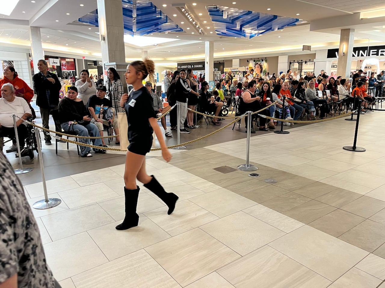 A woman in high heels, a pony tail and a black sweater walks down a runway.