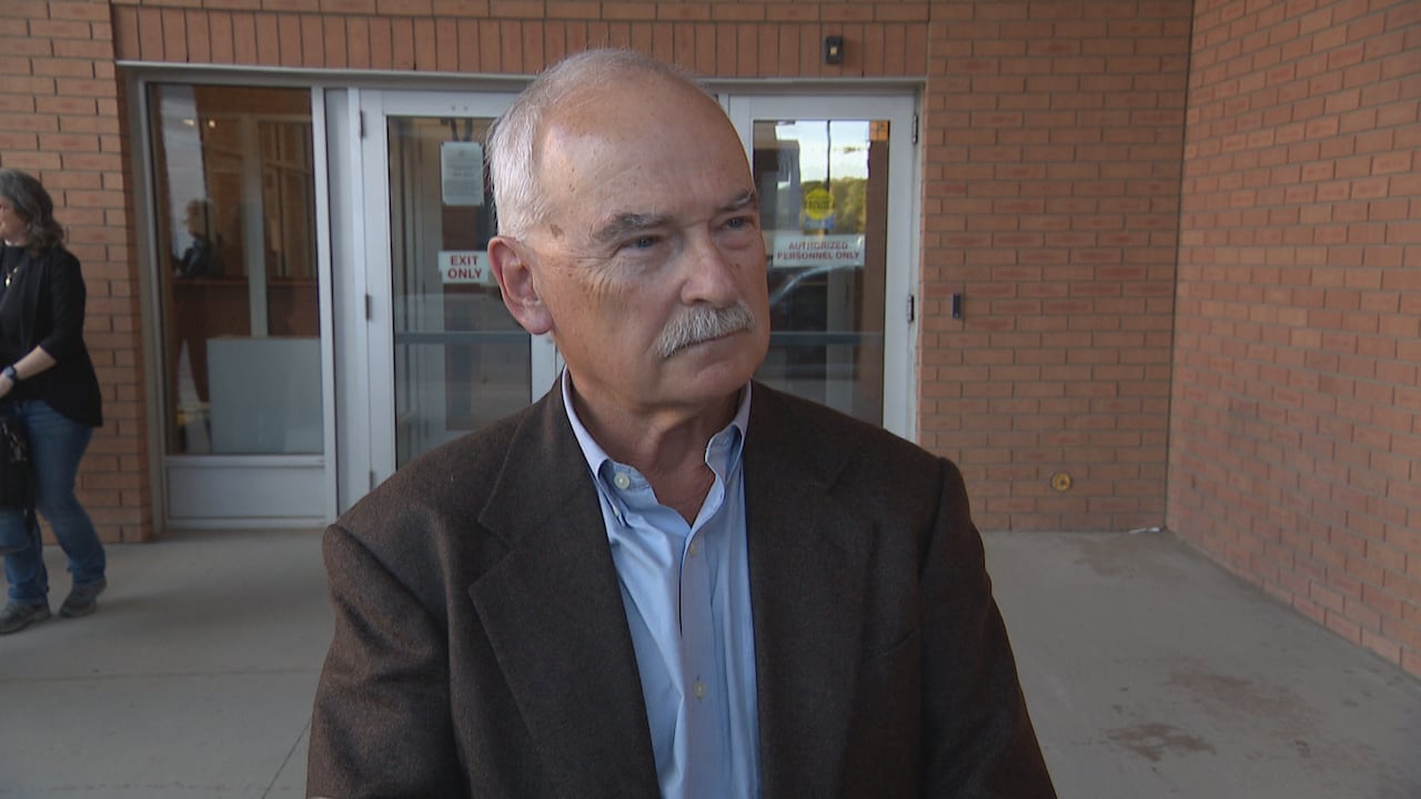 A man looks forward with a serious expression in front of an entrance to a building.