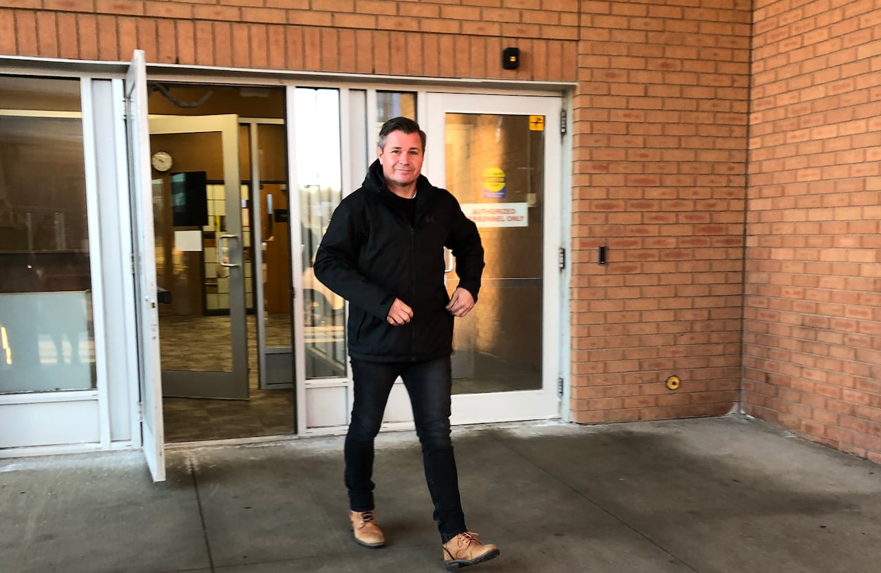 A man in a black sweater and dark pants walks out of a door